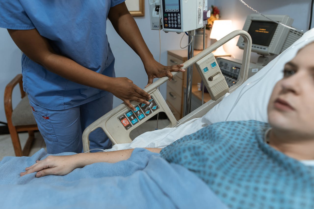 A nurse in scrubs assists a patient in a hospital bed with medical equipment nearby.